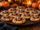 Cookies dorés décorés d’araignées en chocolat aux grands yeux, sur un plateau entouré de petites citrouilles.